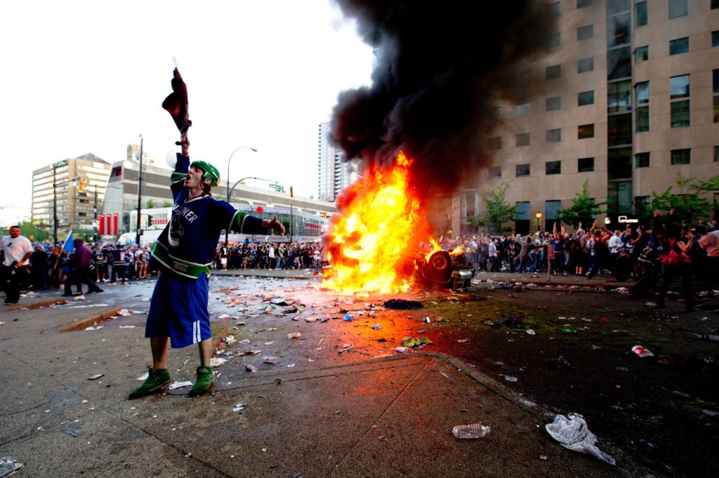 A rioter dressed in a Vancouver Canucks jersey cheers while a car burns, User:Elopde - From Wikimedia Commons, the free media repository, CC BY-SA 3.0, File:Riot in Vancouver.jpg, Created: 15 June 2011